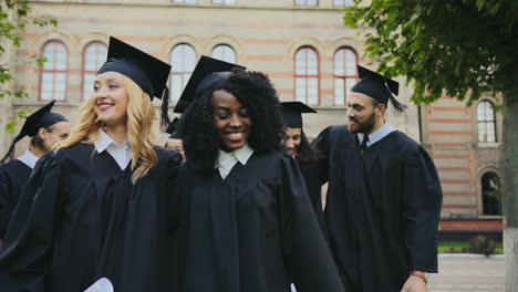 Felices-Sonrientes-Graduados-Multiétnicos-Caminando-Con-Los-Diplomas-En-Las-Manos-Cerca-Del-Edificio-De-La-Universidad-Después-De-La-Ceremonia-De-Graduación