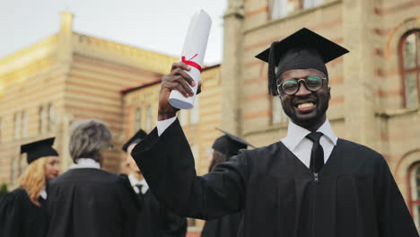 Retrato-De-Un-Joven-Graduado-Afroamericano-Feliz-Posando-Para-La-Cámara-Y-Mostrando-Su-Diploma-Frente-A-La-Universidad