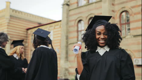 Retrato-De-La-Feliz-Joven-Afroamericana-Graduada-Posando-Para-La-Cámara,-Mostrando-Su-Diploma-Y-Haciendo-Un-Gesto-De-Sí-Frente-A-La-Universidad