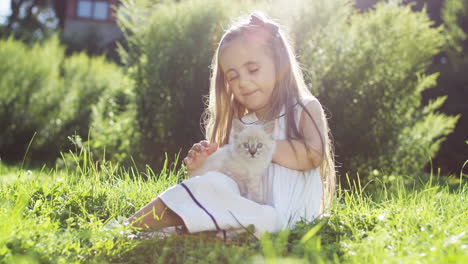 Small-girl-sitting-on-the-green-grass-on-a-sunny-summer-day-and-holding-white-kitty-cat