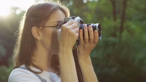 Side-view-of-a-beautiful-young-woman-in-glasses-taking-photos-in-the-park-with-a-old-vintage-photo-camera