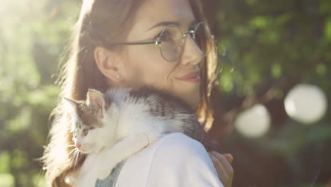 Close-up-view-of-a-caucasian-woman-in-glasses-holding-small-cat-her-shoulder-in-the-park-on-a-summer-day