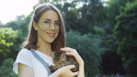 Close-up-view-of-a-caucasian-woman-in-glasses-holding-and-petting-kitty-cat-while-smiling-at-camera-in-the-park-on-a-summer-day