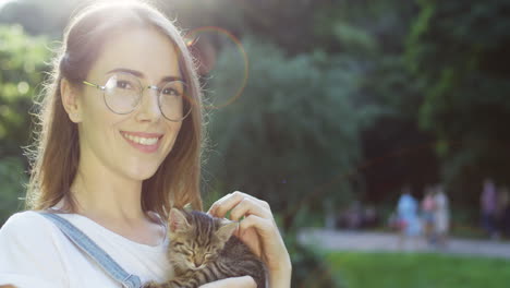 Close-up-view-of-a-caucasian-woman-in-glasses-holding-and-petting-kitty-cat-while-smiling-at-camera-in-the-park-on-a-summer-day