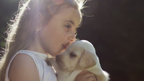 Close-up-view-of-a-caucasian-little-girl-holding-and-kissing-a-small-labrador-puppy-in-the-park-on-a-summer-day