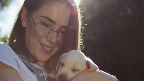 Close-up-view-of-caucasian-young-woman-in-glasses-holding-and-petting-a-labrador-puppy-in-the-park-on-a-summer-day