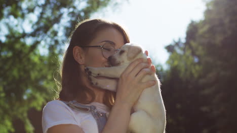 Caucasian-young-woman-in-glasses-holding-and-kissing-a-labrador-puppy-in-the-park-on-a-summer-day