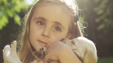 Close-up-view-of-a-caucasian-little-girl-holding-a-small-labrador-puppy-while-looking-at-camera-in-the-park-on-a-summer-day