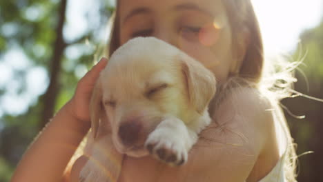 Close-up-view-of-a-caucasian-little-girl-holding-and-petting-a-small-labrador-puppy-while-looking-at-camera-in-the-park-on-a-summer-day