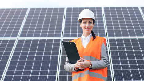 Portrait-of-young-female-engineer-in-special-uniform-and-helmet-holding-a-clipboard-near-solar-panels