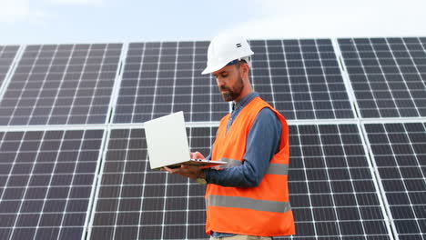 Young-male-engineer-with-helmet-and-vest-near-solar-panels-using-laptop