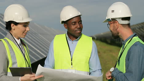 Close-up-view-of-three-multiethnics-farm-solar-engineers-talking-while-looking-at-blueprint-on-solar-plantation