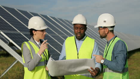 Three-multiethnics-farm-solar-engineers-talking-while-looking-at-blueprint-on-solar-plantation