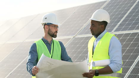 Two-caucasian-and-african-american-farm-solar-engineers-talking-while-looking-at-blueprint-on-solar-plantation
