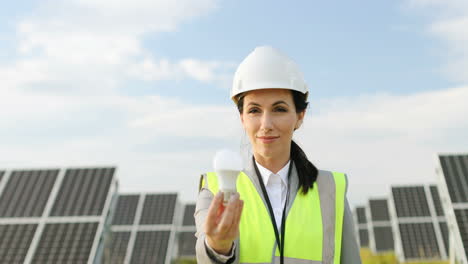 Portrait-of-female-engineer-in-uniform-with-protective-helmet-showing-a-light-bulb-to-the-camera-on-solar-plantation