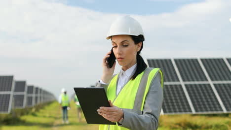 Portrait-of-beautiful-female-engineer-in-protective-helmet-and-uniform-talking-on-the-phone-on-solar-plantation