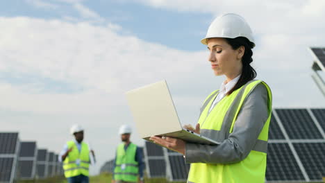 Portrait-of-beautiful-female-engineer-in-protective-helmet-and-uniform-using-laptop-and-looking-around-on-solar-plantation