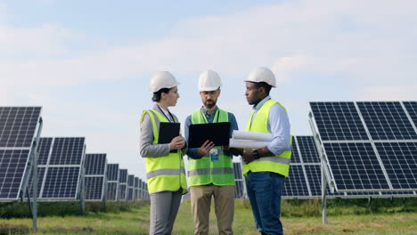 Three-multiethnics-farm-solar-engineers-talking-while-looking-at-blueprint-on-solar-plantation