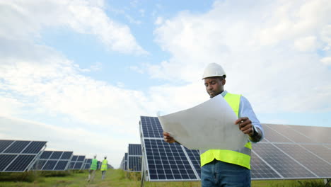 Ansicht-Von-Unten-Eines-Jungen-Afroamerikanischen-Ingenieurs,-Der-Einen-Solarplan-Auf-Einer-Solarplantage-Hält-Und-Betrachtet