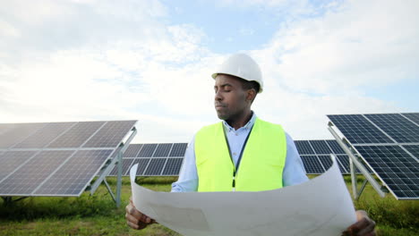 Vorderansicht-Eines-Jungen-Afroamerikanischen-Ingenieurs,-Der-Einen-Solarplan-Hält-Und-Sich-Auf-Einer-Solarplantage-Umsieht