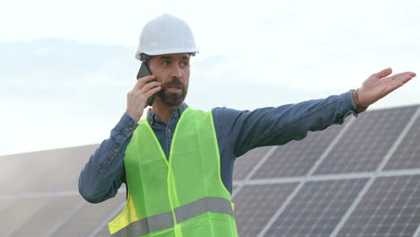Caucasian-man-in-special-uniform-and-protective-helmet-and-giving-instructions-and-talking-on-the-phone-on-solar-plantation