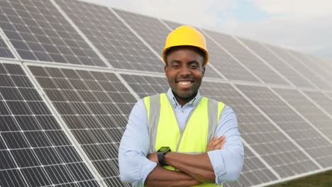 Vista-De-Cerca-De-Un-Joven-Ingeniero-Afroamericano-Con-Casco-Protector-Sonriendo-A-La-Cámara-Con-Los-Brazos-Cruzados-En-Una-Plantación-Solar