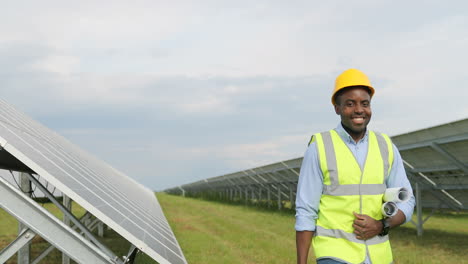 Happy-african-american-engineer-walking-and-holding-rolls-of-paper-with-diagrams-and-tool-box-on-solar-plantation