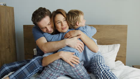 Rear-of-cute-little-boy-hugging-his-mother-and-father-while-they-are-sitting-on-the-bed-in-the-morning