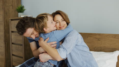 Cute-little-boy-hugging-his-mother-and-father-while-they-are-sitting-on-the-bed-in-the-morning