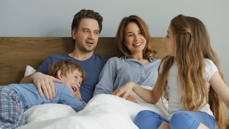 Kaukasische-Glückliche-Freundliche-Eltern-Mit-Ihrem-Sohn-Und-Ihrer-Tochter,-Die-Morgens-Auf-Dem-Bett-Sitzen