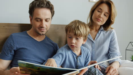 Young-mother-and-father-sitting-on-the-bed-in-the-morning-with-their-little-son-and-reading-an-interesting-book