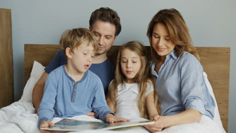 Young-mother-and-father-sitting-on-the-bed-in-the-morning-with-their-little-son-and-daughter,-they-are-reading-an-interesting-book
