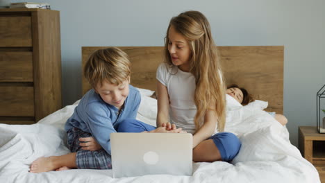 Süße-Kleine-Kinder,-Die-Vor-Der-Kamera-Auf-Dem-Bett-Liegen-Und-Einen-Laptop-Benutzen,-Während-Ihre-Eltern-Morgens-Hinter-Ihnen-Liegen