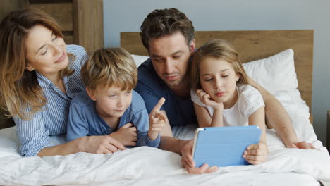 Happy-smiled-family-of-mother,-father,-daughter-and-son-lying-on-the-bed-in-the-morning-and-having-video-call-on-a-tablet