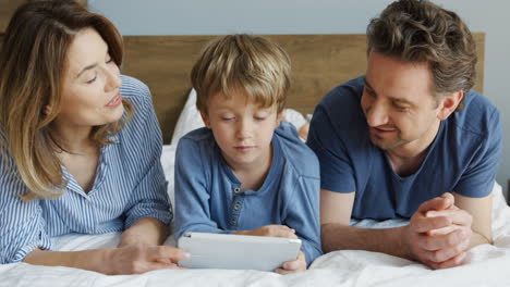 Close-up-view-of-a-little-boy-and-his-parents-lying-on-the-bed-and-watching-something-on-the-tablet