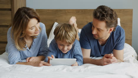 Nahaufnahme-Eines-Kleinen-Jungen-Und-Seiner-Eltern,-Die-Auf-Dem-Bett-Liegen-Und-Etwas-Auf-Dem-Tablet-Beobachten