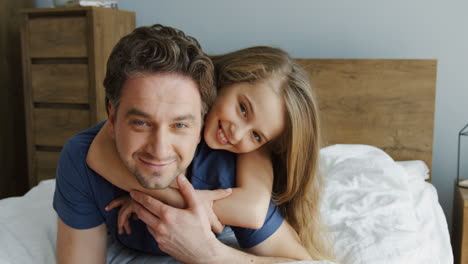 Close-up-view-of-happy-father-and-his-blonde-daughter-hugging-and-lying-on-the-bed-while-looking-at-camera