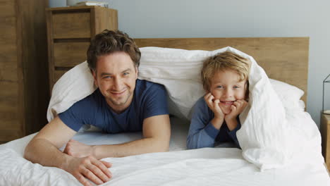 Cheerful-funny-father-and-little-son-laughing-and-looking-out-of-the-blanket-while-lying-on-the-bed-in-the-morning