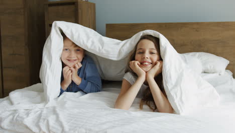 Cute-funny-little-sister-and-brother-smiling-and-looking-out-of-the-blanket-while-lying-on-the-bed-in-the-morning