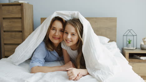 Hübsche-Kaukasische-Mutter-Und-Ihre-Süße-Tochter-Lächeln-Und-Schauen-Aus-Der-Decke,-Während-Sie-Morgens-Auf-Dem-Bett-Liegen