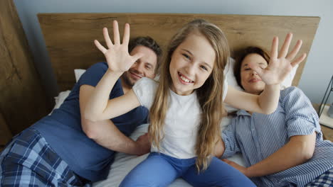 Top-view-of-a-blonde-girl-sitting-on-the-bed-and-waving-her-hands-to-the-camera-between-her-parents-who-are-lying