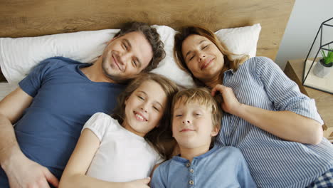 Top-view-of-happy-parents-with-their-kids-lying-on-the-bed,-hugging-and-smiling-at-camera-in-the-morning