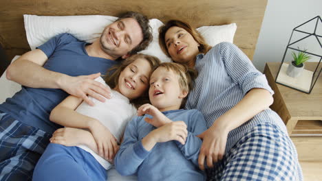 Top-view-of-happy-parents-with-their-kids-lying-on-the-bed-and-hugging-while-they-talking