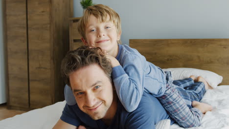 Close-up-view-of-happy-father-and-his-cute-little-son-hugging-him-from-back,-while-they-lying-on-the-bed-and-looking-at-camera-in-the-morning