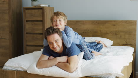 Happy-father-and-his-cute-little-son-hugging-him-from-back,-while-they-lying-on-the-bed-and-looking-at-camera-in-the-morning