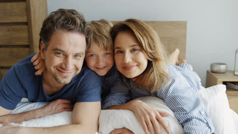 Close-up-view-of-happy-father-and-mother-with-his-cute-little-son-hugging-while-they-lying-on-the-bed-and-looking-at-camera-in-the-morning