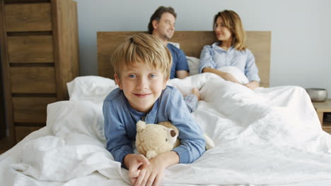 Cute-small-boy-lying-with-a-teddy-bear-and-smiling-at-camera,-his-parents-are-lying-under-the-blanket-on-the-bed-behind-him