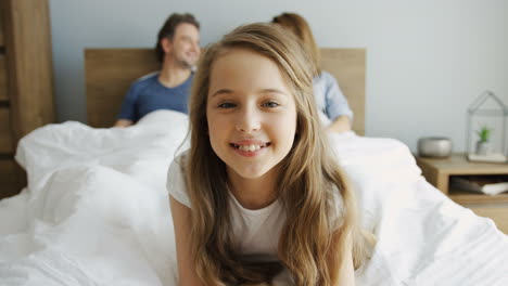 Cute-little-girl-lying-and-smiling-at-camera,-her-parents-are-lying-under-the-blanket-on-the-bed-behind-her