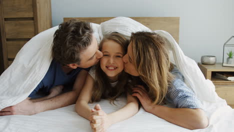 Cheerful-parents-looking-out-of-the-blanket-and-kissing-her-daughter-while-they-are-lying-on-the-bed-and-smiling-at-camera