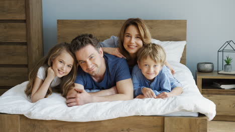 Cheerful-parents-and-their-sons-lying-on-the-bed-and-smiling-at-camera-in-the-morning